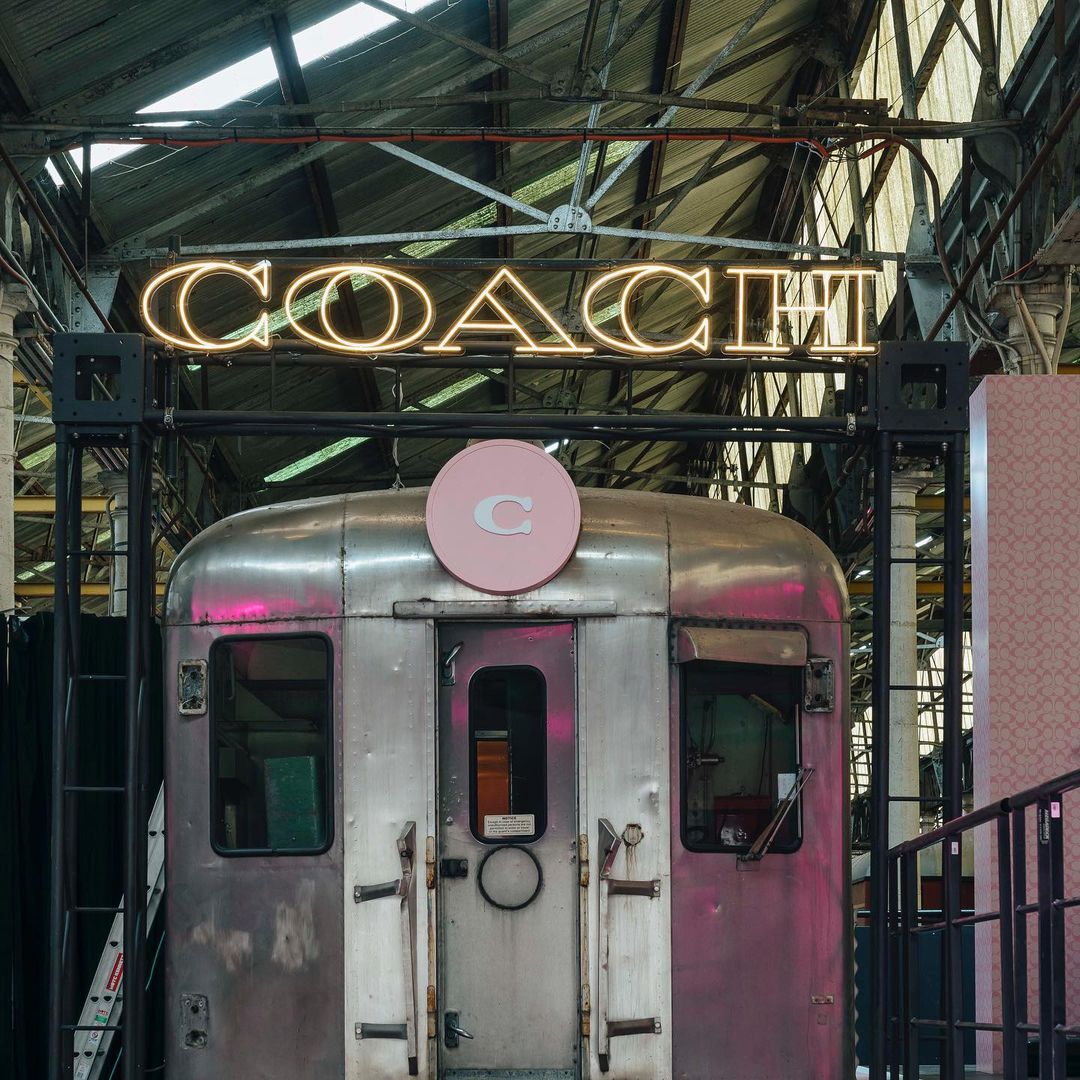 Vintage Coach subway car with neon signage. The train's metallic exterior and pink accents contrast with industrial interior and brand logo.