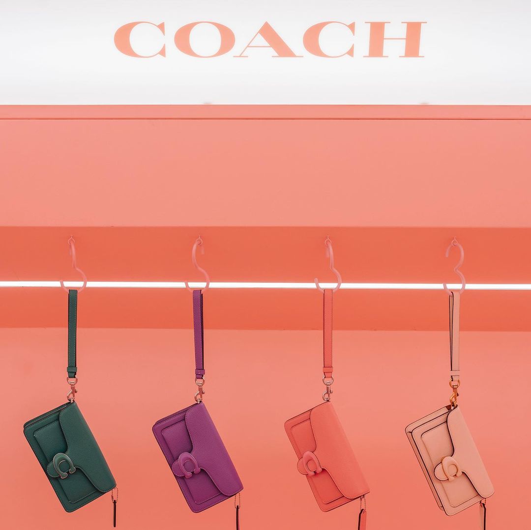 Four Coach purses in green, purple, peach, and pink hang on hooks against a matching pink wall with the Coach logo above.