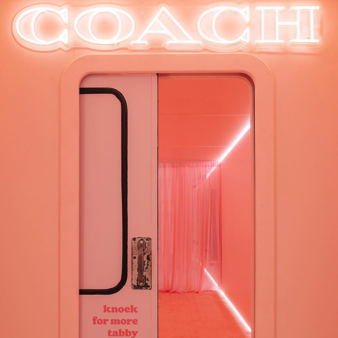 Coach neon sign above a pink door leading to a room with matching pink decor and a light fixture. Fashion-forward interior design.