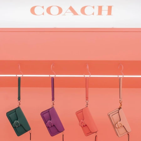 Four Coach leather wristlet bags in green, purple, peach, and pink hanging against a coral pink wall