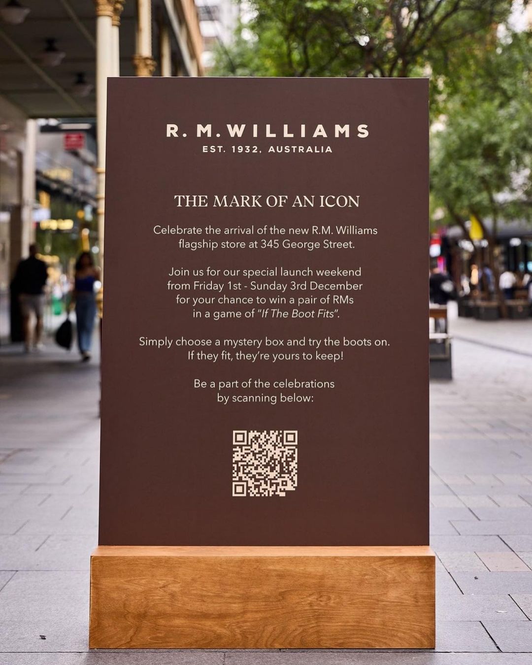 R.M. Williams flagship store advertisement on a brown sign with event details, QR code for scanning, and a wooden base on a sidewalk.