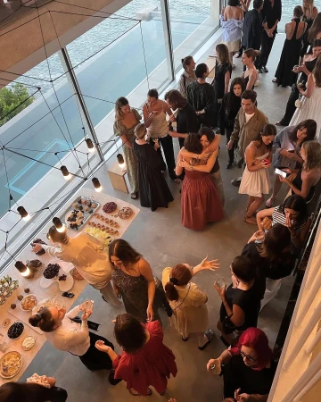 Indoor social gathering with many people in a naturally lit hall with large windows, enjoying a celebration with tables of food.