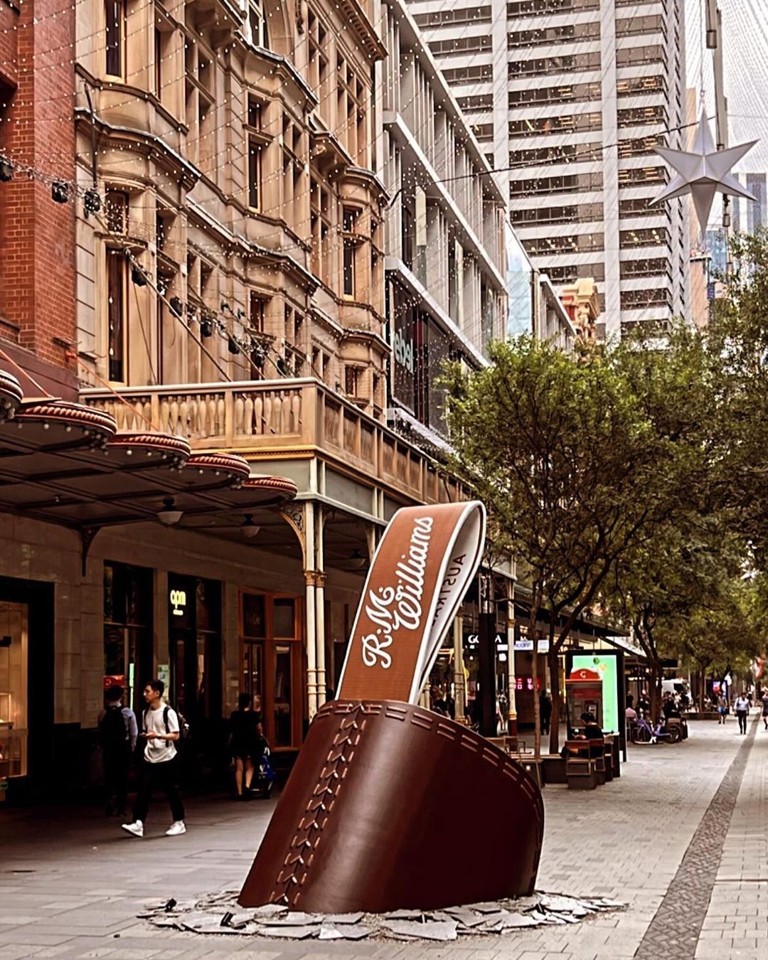Outdoor shot of an R.M. Williams boot sculpture on a brick-paved city street lined with buildings, trees, and pedestrians.
