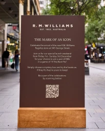 Brown promotional sign on a wooden base placed on a paved outdoor walkway in Sydney, featuring brand text and a QR code.