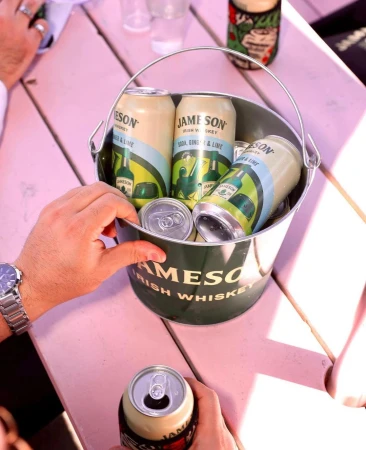 Overhead shot of a bucket filled with Jameson Irish Whiskey canned cocktails on a pink wooden table, held by a person wearing a watch.