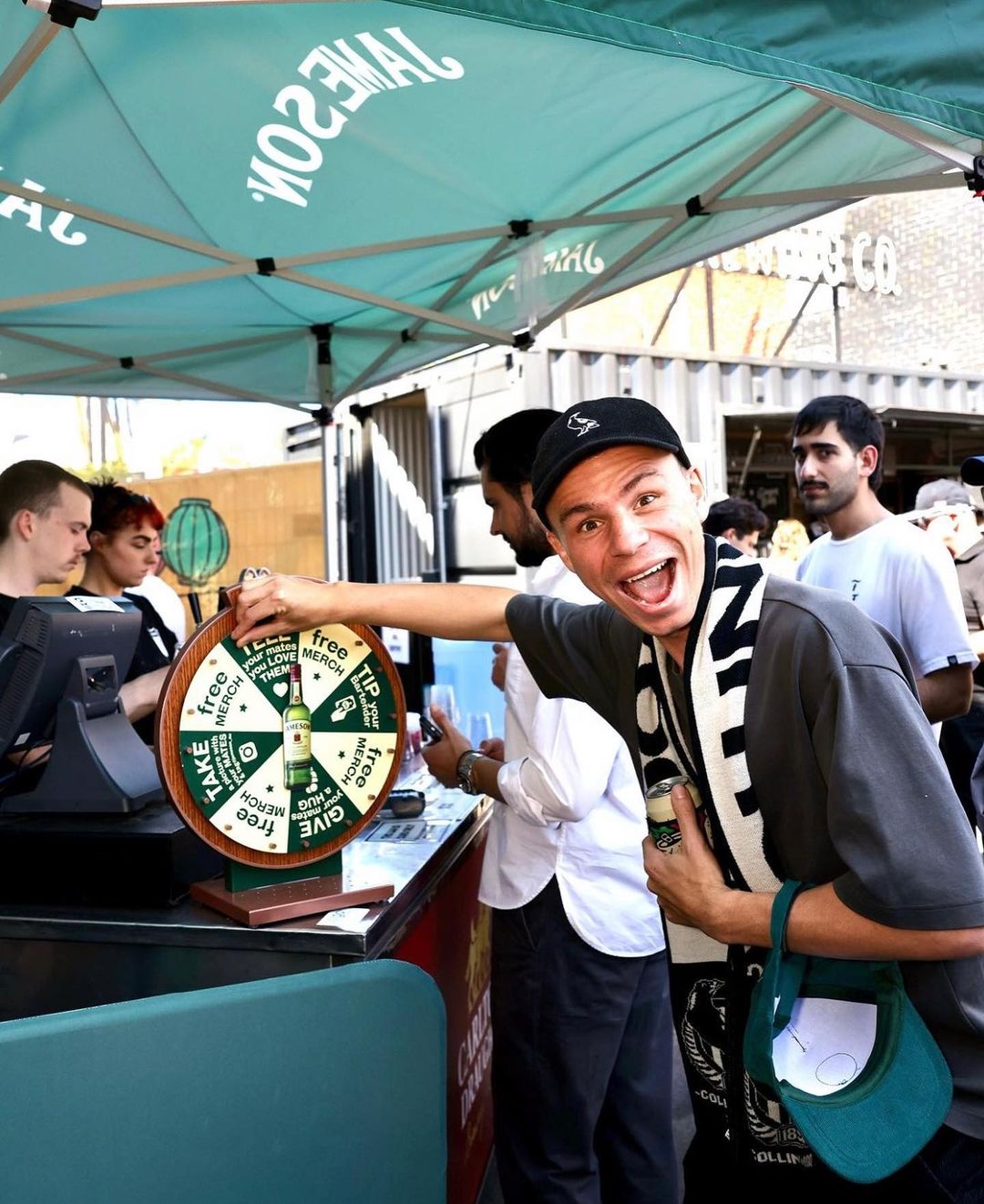 Enthusiastic man spinning a prize wheel at an outdoor Jameson Irish Whiskey promotional event, with blurred people in the background.