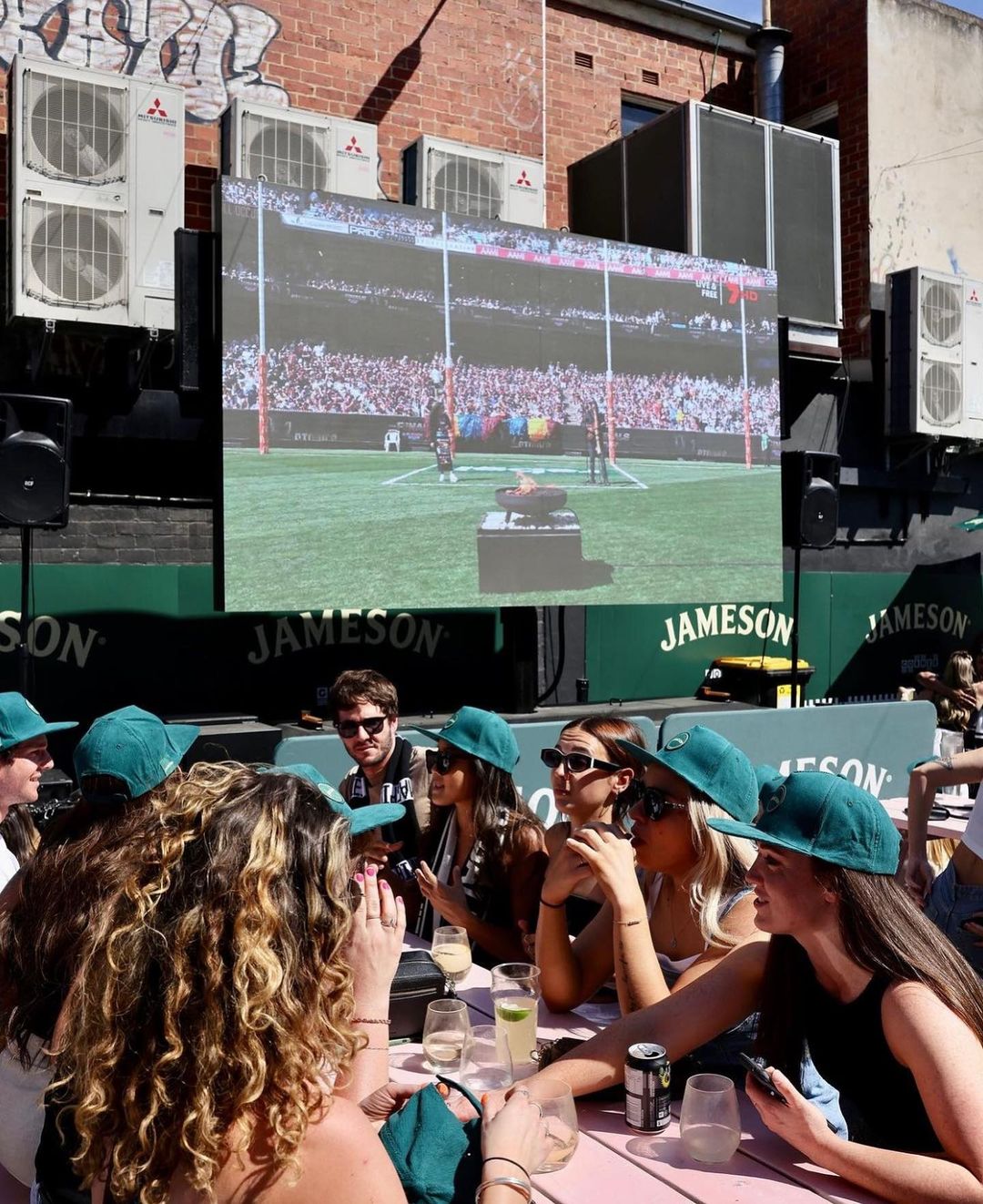 Group of people watching a sports game on a large screen in an outdoor setting, socializing with drinks and wearing matching green hats.