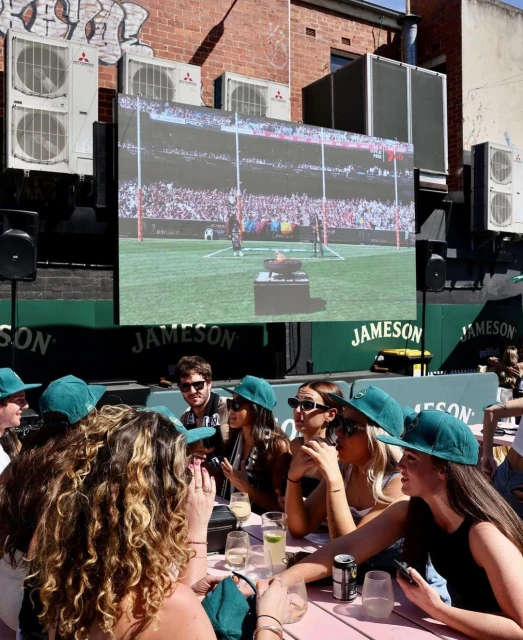 Group of people watching a sports game on a large screen in an outdoor setting, socializing with drinks and wearing matching green hats.