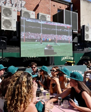 Group of people watching a sports game on a large screen in an outdoor setting, socializing with drinks and wearing matching green hats.