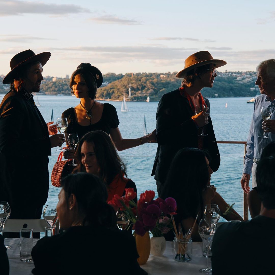 Group of people in formal attire gather on a terrace overlooking a bay, enjoying drinks and conversation at an outdoor event.