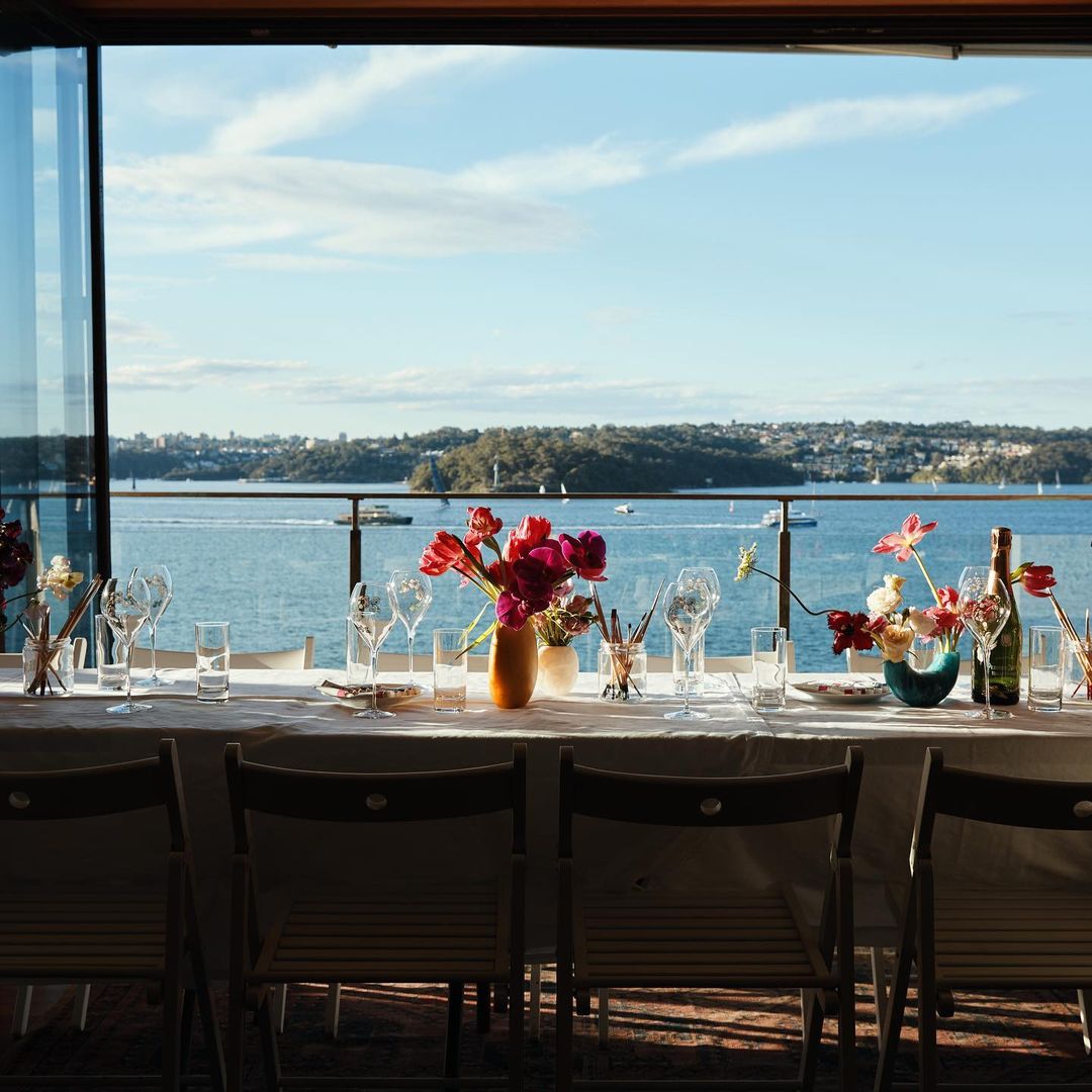 Formal dining table setting with flower arrangements and glassware, presented with a scenic water and shoreline backdrop, bathed in natural light.