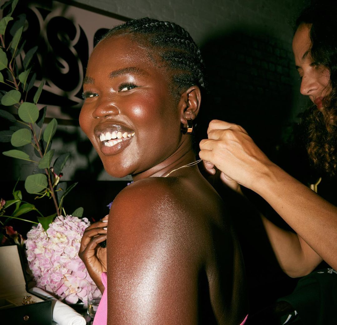 A smiling Black woman has a necklace being put on her by another person in a room decorated with floral arrangements and foliage.