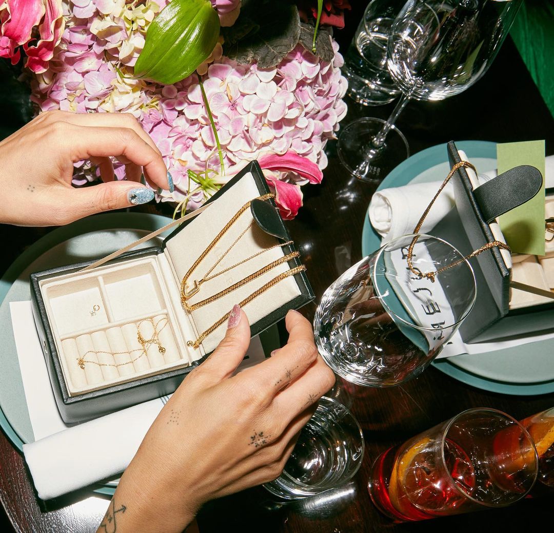 Overhead shot of hands opening a black jewelry box with gold necklaces, surrounded by drinks, glassware, plates, and pink flowers.