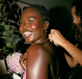 Smiling black woman wearing gold hoop earrings and pink top while another person adjusts her necklace