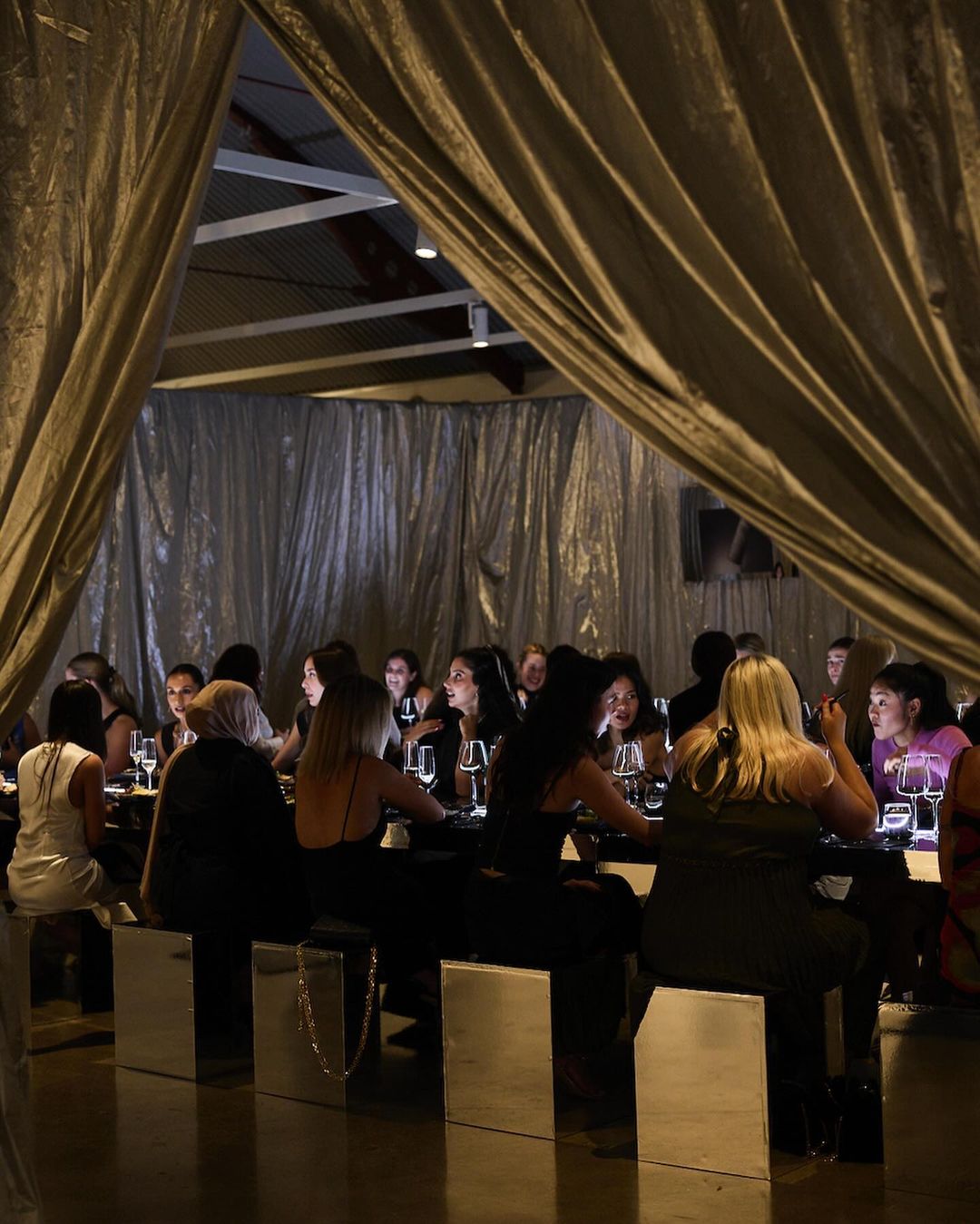 A group of people are seated at a long table in an indoor setting with dramatic lighting, silver chairs and hanging metallic drapery.
