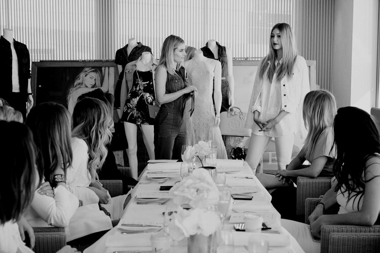 Black and white image of a fashion presentation with several women seated around a table and mannequins displayed in the background.
