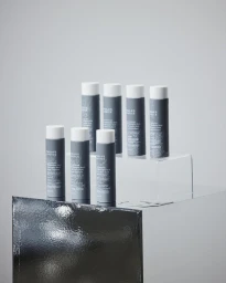 Seven dark grey cylindrical skincare bottles with white caps arranged on two tiered reflective display blocks against a light grey background.