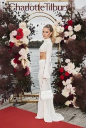 Woman in a long white two-piece dress holding a white handbag standing on a red carpet in front of a floral archway in Sydney.