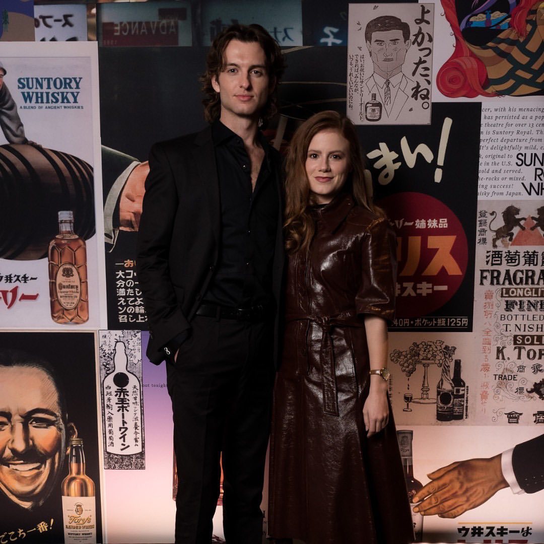 A man and a woman pose together in front of a wall covered in Japanese whisky advertisements, creating a retro and stylish scene.