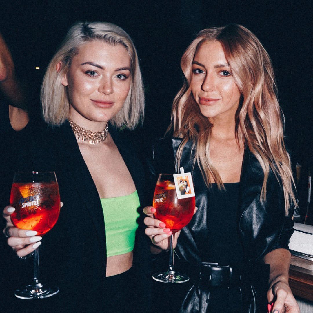 Two blond women pose indoors, holding glasses with red drinks, wearing black tops, with dark background in a medium eye-level shot.
