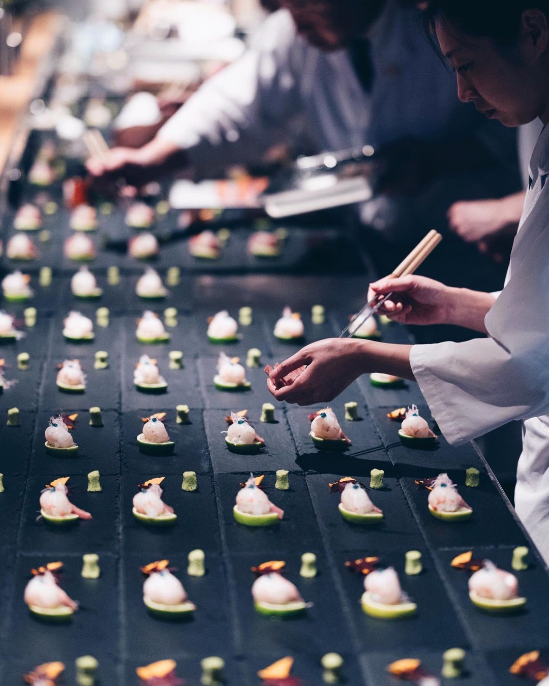 Two chefs prepare a multitude of appetizers arranged in rows, featuring fish, green citrus, and wasabi, in an indoor restaurant setting.