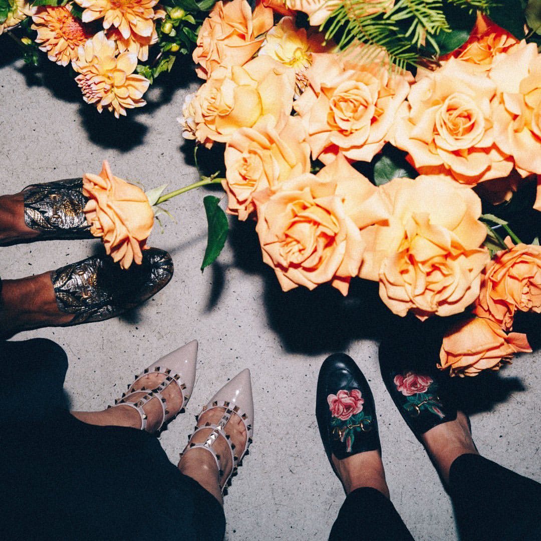Overhead shot of four pairs of legs and feet wearing different fashionable shoes, surrounded by peach-colored roses and dahlias.