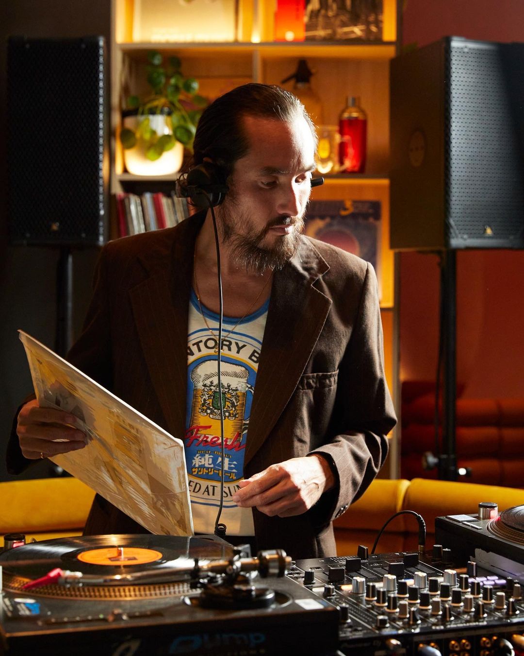 DJ mixing music in dimly lit venue. Adult male with beard wearing headphones and blazer. Turntables, record albums, sound equipment visible.