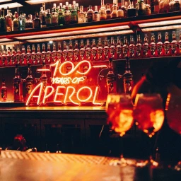 Illuminated Aperol neon sign reading 100 years of Aperol behind a bar shelf stocked with liquor bottles.