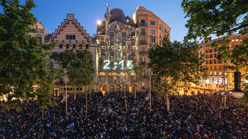 Wide shot of a crowd of people gathering at night to view a building with a digital display near Plaça de Catalunya, Barcelona.