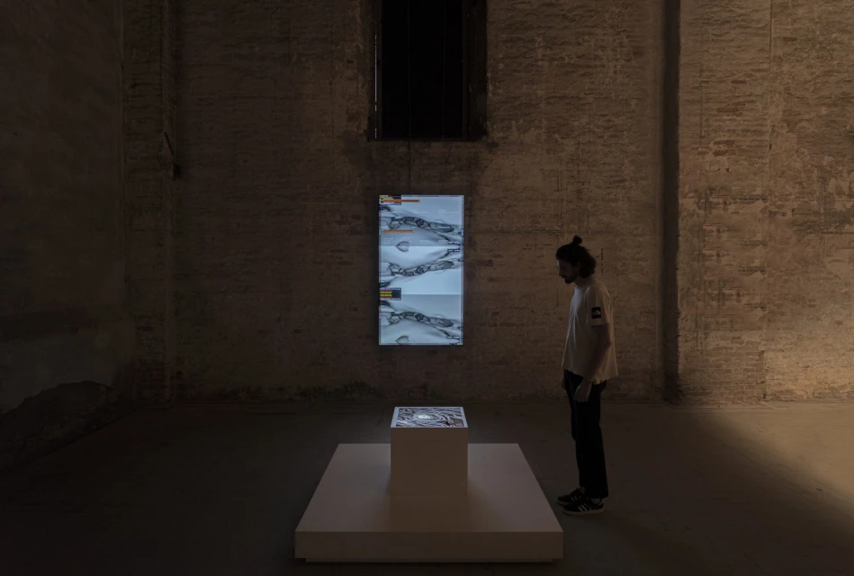 Man stands in dimly lit room examining an illuminated display on a pedestal and a hanging screen showing abstract imagery.