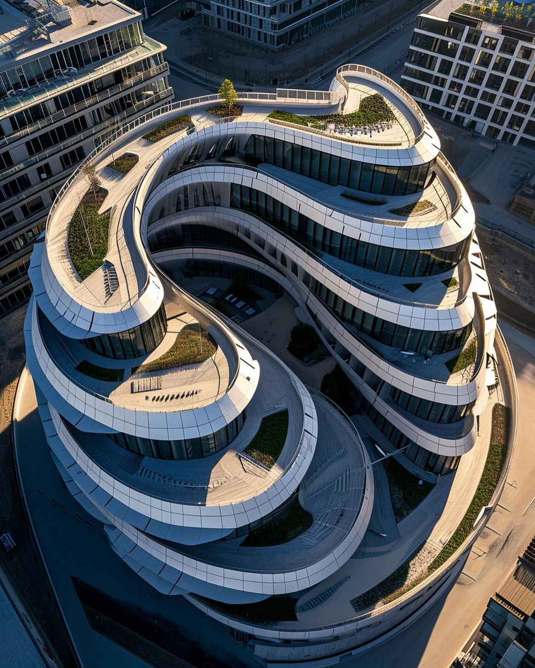 High-angle view captures a modern, curvilinear office building with tiered, landscaped roofs, set in an urban environment with nearby buildings.
