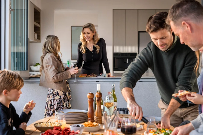 Family gathering around wooden table indoors with food, wine, and drinks in modern home setting with natural light and cheerful atmosphere.