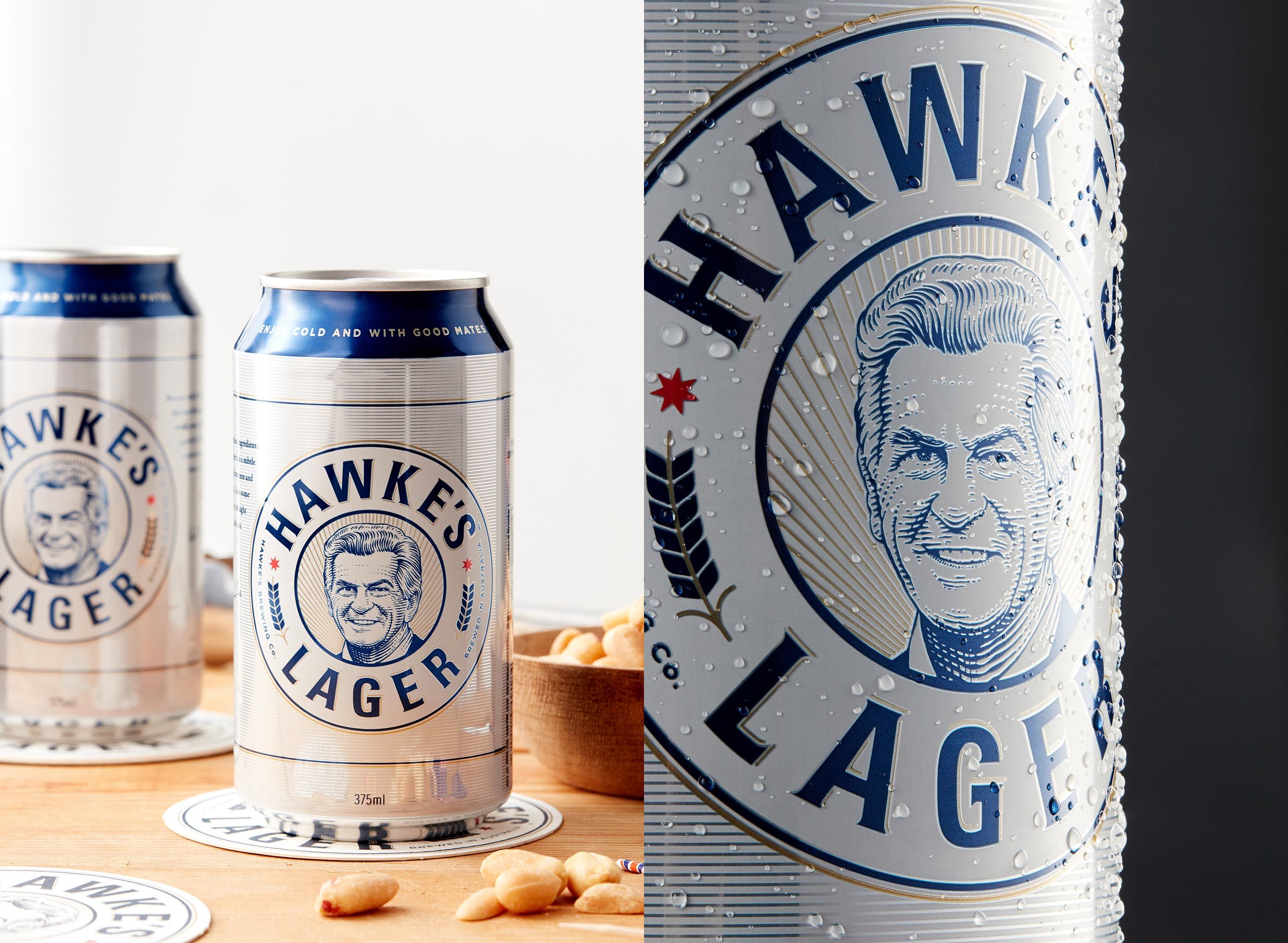 Two silver beer cans of Hawke's Lager on a wooden table with peanuts. Close-up of can shows water droplets. Product photography.
