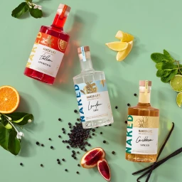 Three bottles of non-alcoholic spirits surrounded by fresh citrus slices, juniper berries, and mint leaves on a green surface.