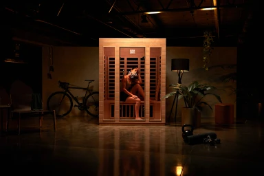 A woman seated inside a wooden sauna cabin in a dimly lit industrial studio space styled by Kalliroy Edlin.