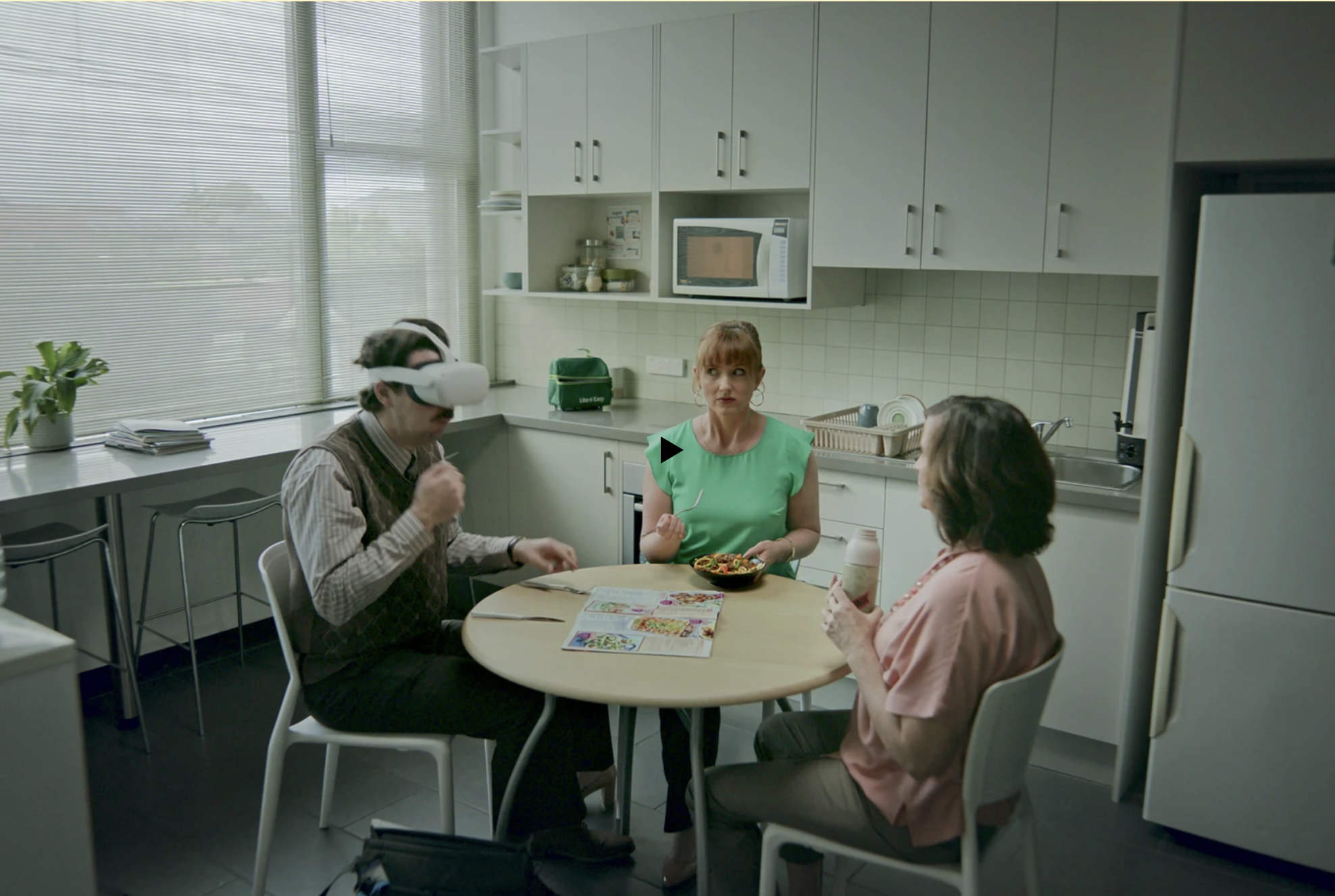 Three adults at a round table in a bright kitchen; one wearing VR headset, another holding a bowl of food, and another holding a bottle.