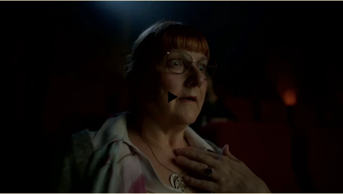 A medium shot of a mature woman with a surprised expression, wearing glasses, a patterned shirt, and a necklace in a dark theater setting.