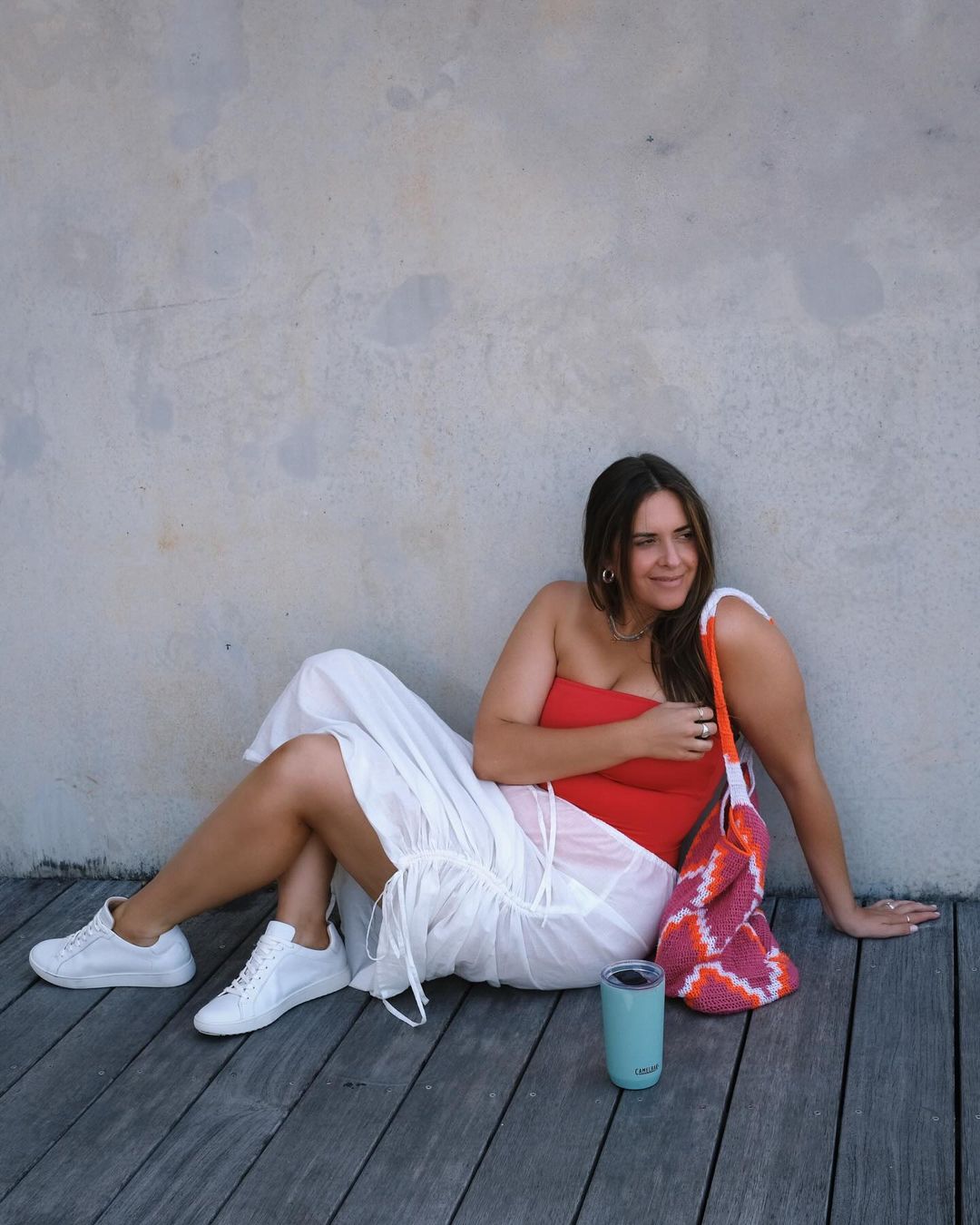 An adult woman with long brown hair sits on a wood floor, wearing a red top and white skirt, with a colorful bag and drink container.