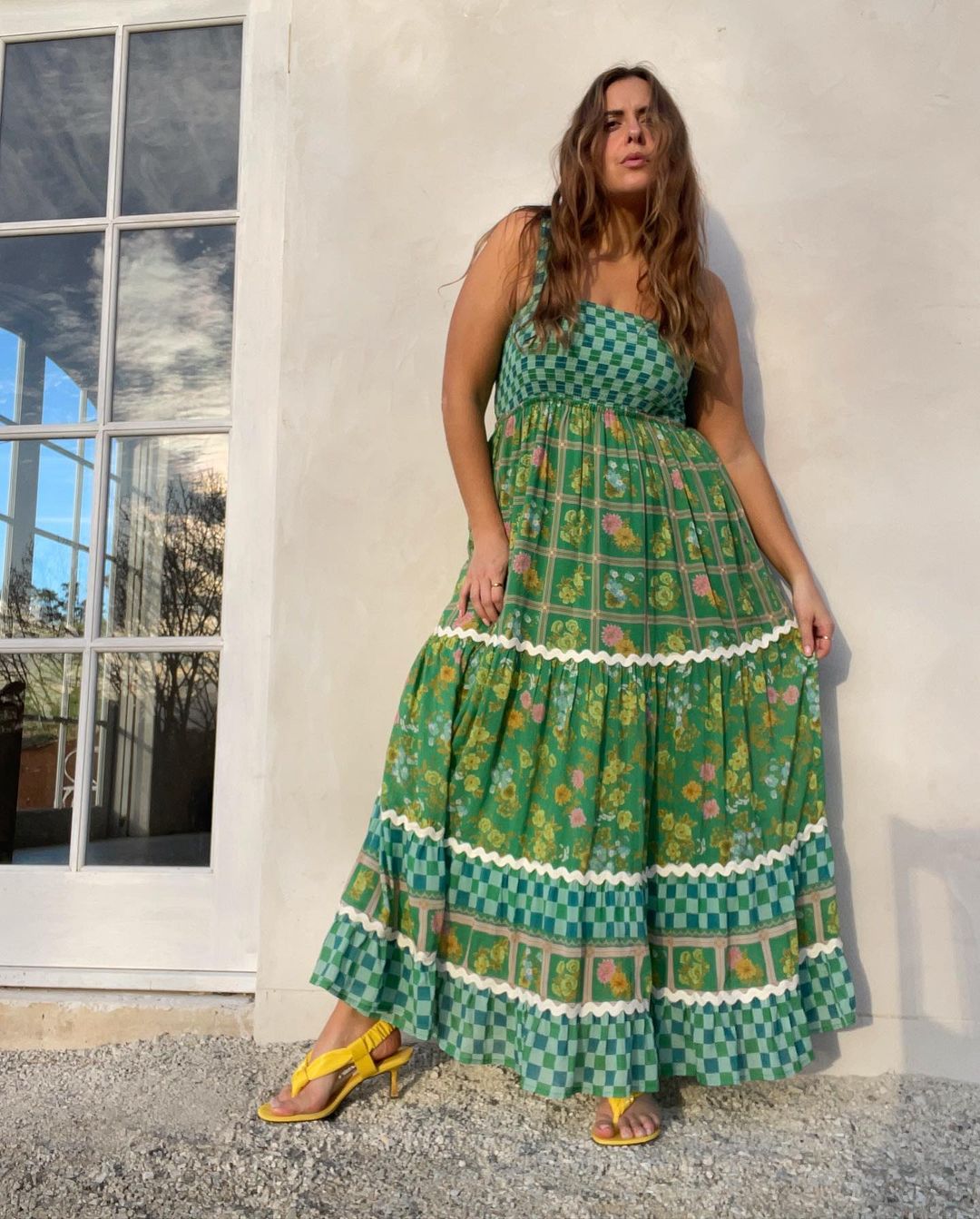 Woman in a green floral maxi dress standing in front of a white building with large windows, wearing yellow heeled sandals.