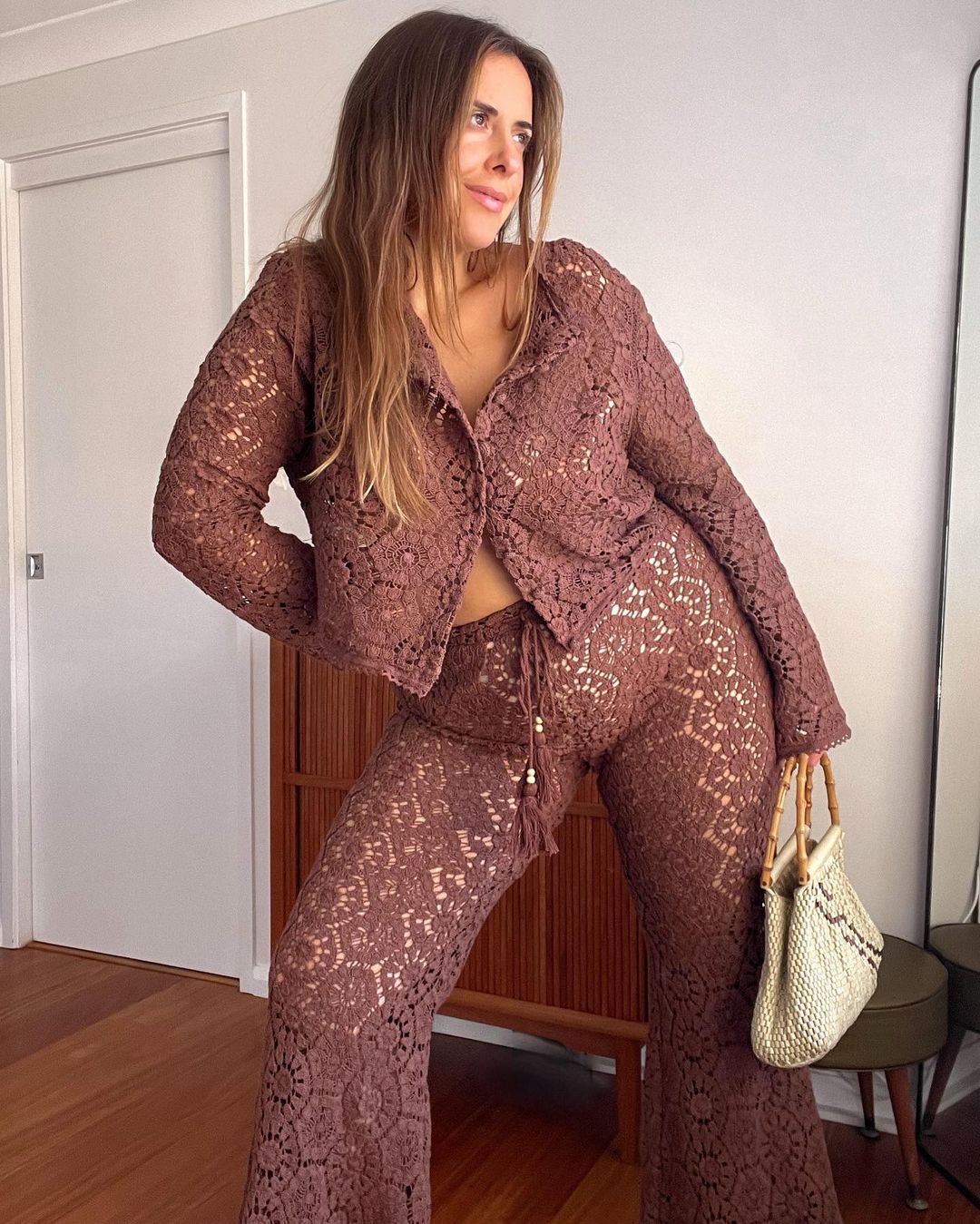 Woman in a brown lace matching set stands indoors with a neutral background. She holds a woven handbag with bamboo handles.