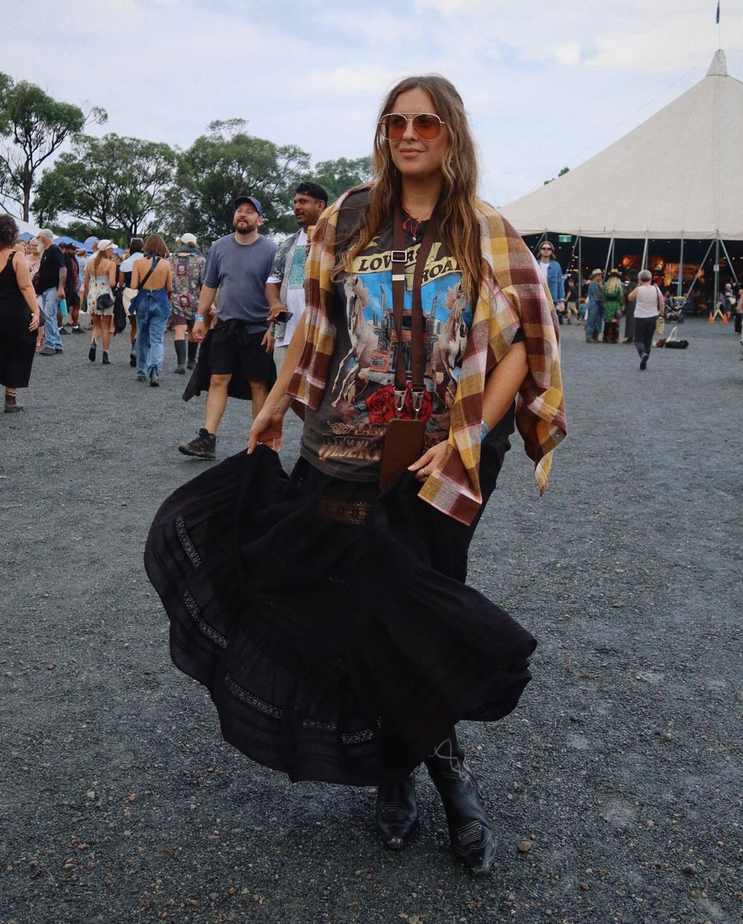 Woman in vintage t-shirt and black skirt at an outdoor festival, accessorized with sunglasses and boots, with a crowd of people.