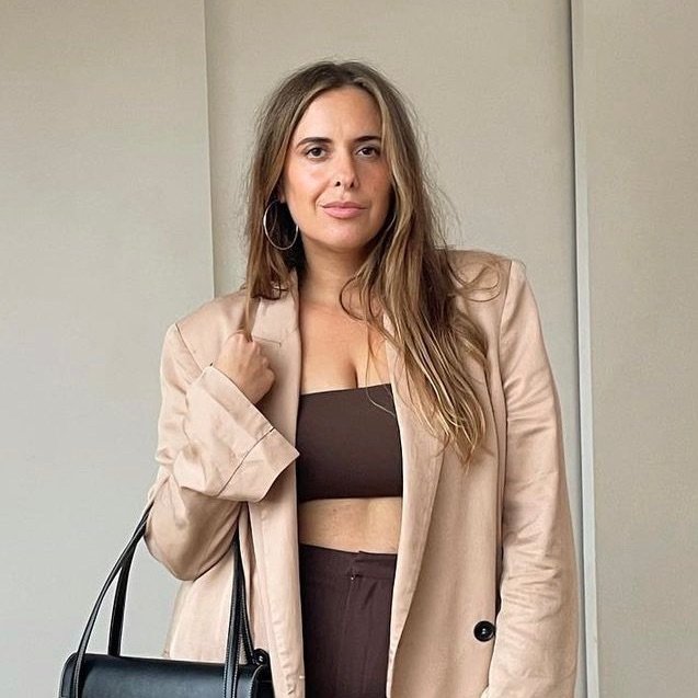 A woman poses indoors wearing a beige blazer, brown crop top and pants, and carrying a black purse, showcasing a casual fashion style.