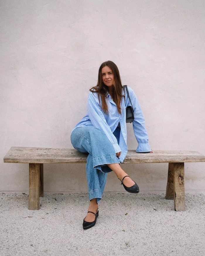 Woman wearing blue striped shirt and wide-leg jeans seated on rustic wooden bench outdoors