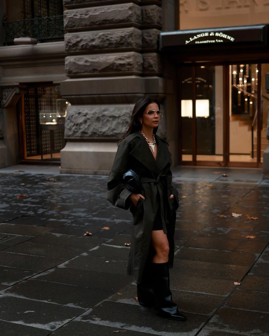 A woman in a dark coat and high black boots walks on a city sidewalk with a purse, in front of a building with exterior lighting.