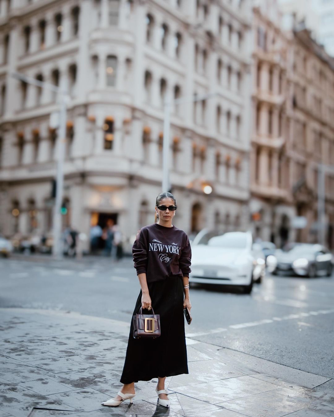 Adult woman in a sweatshirt and skirt standing on a wet street with city buildings, a handbag, sunglasses, and white heeled shoes.