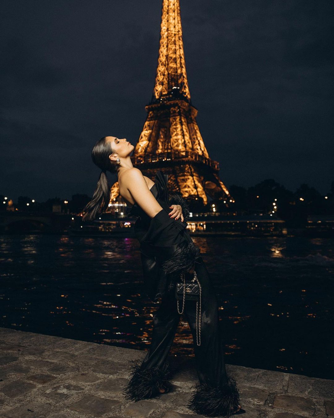 Woman in evening wear poses against the backdrop of the illuminated Eiffel Tower at night, with the Seine River in view.