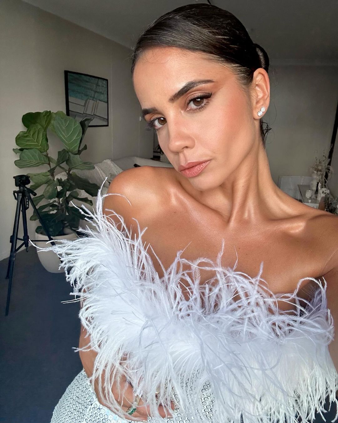 Close-up portrait of a woman with brown hair and brown eyes wearing a white feather top posing indoors with a plant in the background.