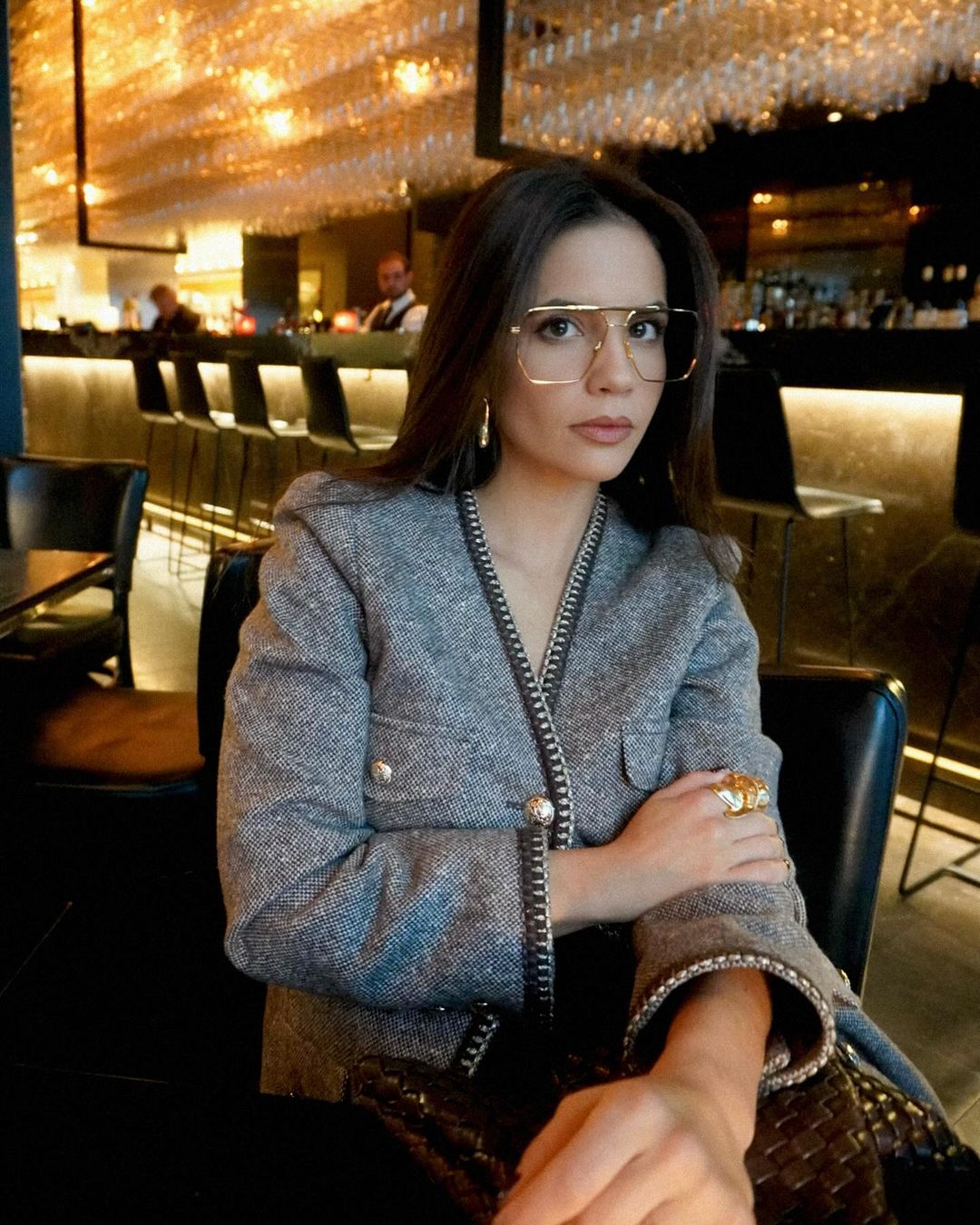 Attractive brunette woman in stylish grey jacket and retro eyeglasses sitting in upscale bar, relaxing with elegant gold jewelry.