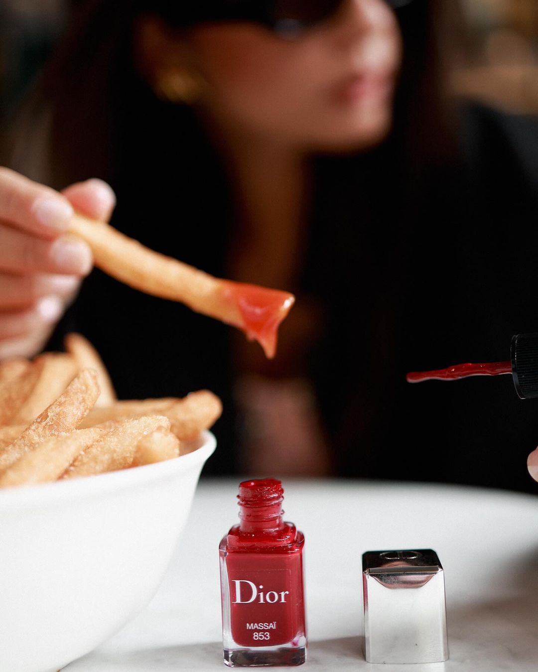 Close-up of a woman with sunglasses dipping a French fry with ketchup near Dior nail polish, creating a fusion of beauty and indulgence.