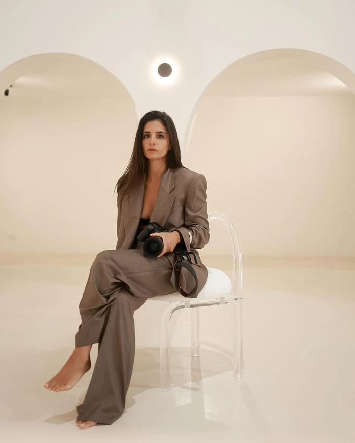 Woman in brown suit sitting on clear acrylic chair holding camera